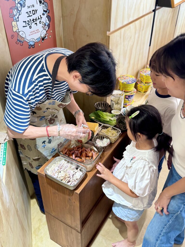 2026년 8월 29일 MR.팬지 작가 북토크와 라면 파티 — 앞치마를 두른 작가가 재료 상자 앞에서 어린이에게 라면 재료를 덜어 주고 있어요