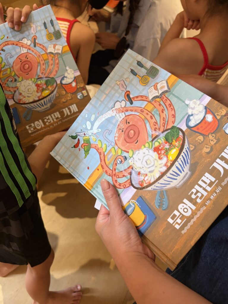2026년 8월 29일 MR.팬지 작가 북토크와 라면 파티 — 어린이들이 『문어 라면 가게』 그림책을 들고 표지를 함께 보고 있어요