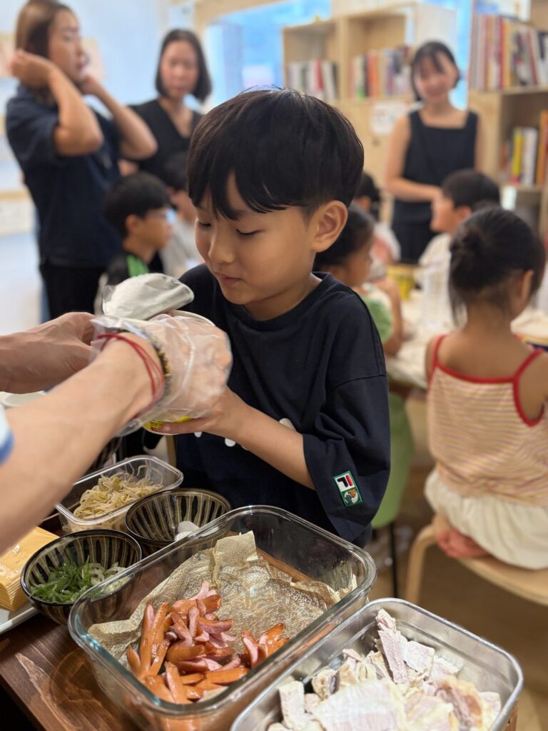 2026년 8월 29일 MR.팬지 작가 북토크와 라면 파티 — 어린이가 국자를 건네받아 자기 그릇에 재료를 담고 있어요
