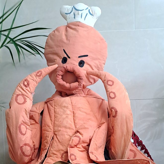 Mr. 팬지 작가. 사람 크기만 한 주황색 문어 인형 옷을 입고, 하얀 요리사 모자를 쓴 채 다리 두 개를 얼굴 옆에 올리고 앉아 있어요.