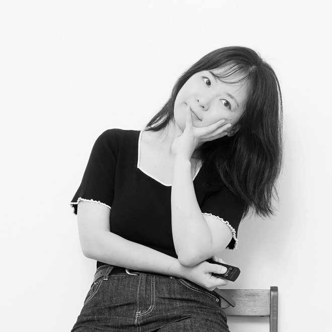 양소이 작가. 흰 벽 앞 나무 의자에 기대어 한 손으로 턱을 괴고 웃고 있는 흑백 사진이에요.